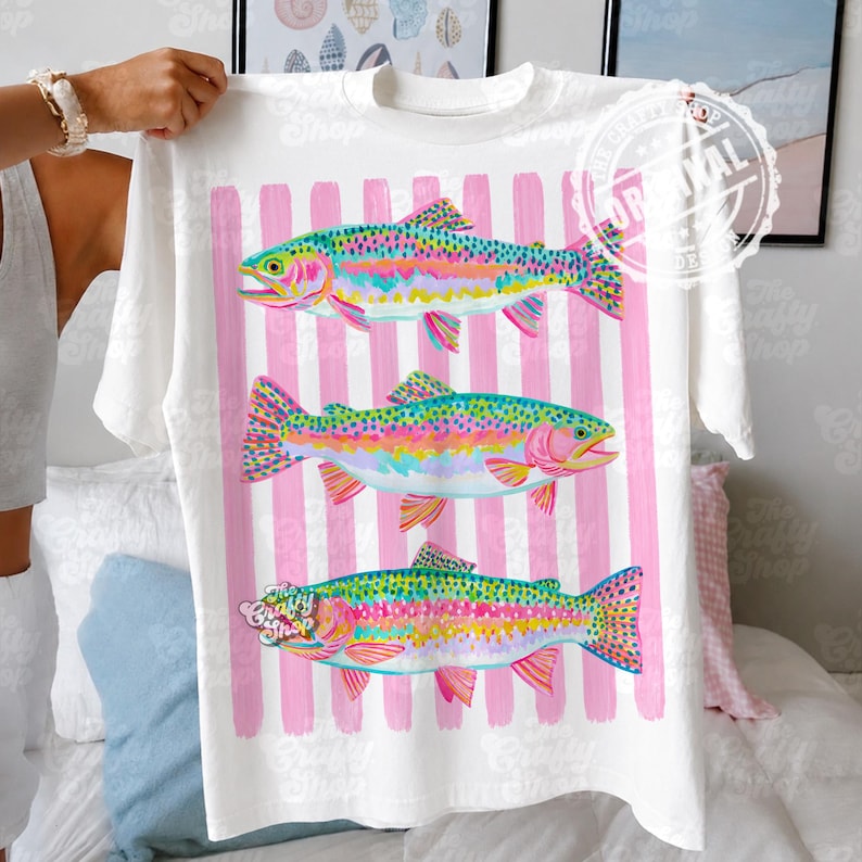 May include: White t-shirt with three vibrant rainbow trout designs. The fish are set against a backdrop of pink vertical stripes. The text "The Crafty Shop" is printed on the shirt.