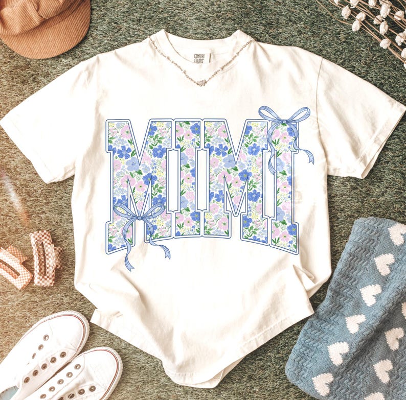 May include: White t-shirt with the word "MIMI" in large, floral-filled letters. The letters are outlined in blue and adorned with blue bows. The floral pattern includes blue and pink flowers. A silver necklace is visible.