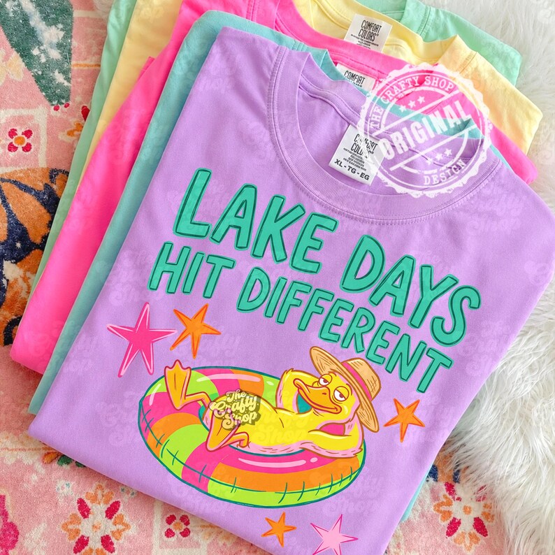 May include: A selection of t-shirts in shades of pink, teal, yellow, and purple. The front shirt is lilac with the text "LAKE DAYS HIT DIFFERENT" and a graphic of a duck in a floatie. The shirts have the "Comfort Colors" brand tag.