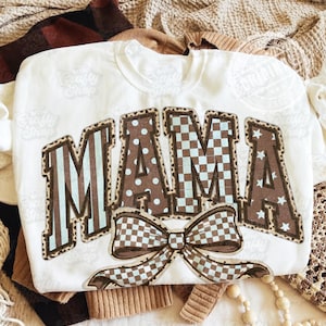 Pode incluir: Camisola branca com a palavra "MAMA" em letras castanhas com vários padrões, incluindo riscas, bolinhas e estrelas. Um grande laço com um padrão xadrez está abaixo da palavra. O logótipo The Crafty Shop é visível.