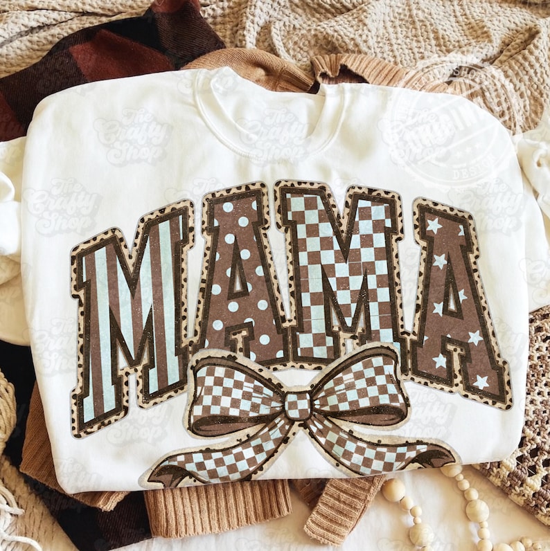 May include: White sweatshirt featuring the word "MAMA" in brown letters with diverse patterns, including stripes, polka dots, and stars. A large bow with a checkered pattern is below the word. The Crafty Shop logo is visible.
