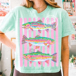 May include: A pale green t-shirt with three rainbow trout illustrations. The fish are depicted in vibrant colours against a backdrop of pink stripes. The tee has a relaxed fit and short sleeves.