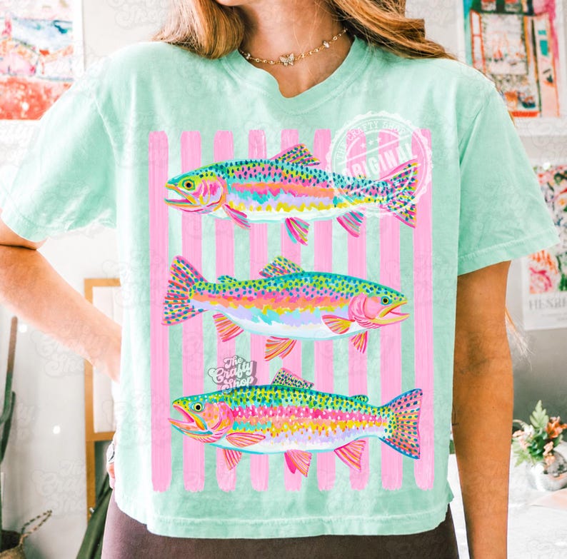 May include: A pale green t-shirt with three rainbow trout illustrations. The fish are depicted in vibrant colours against a backdrop of pink stripes. The tee has a relaxed fit and short sleeves.