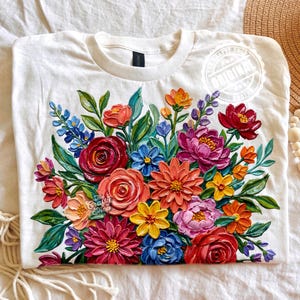 Pode incluir: Camiseta branca com um design vibrante de buquê floral. O buquê inclui flores vermelhas, rosas, laranjas, amarelas e azuis com folhas verdes. A camisa tem gola redonda e mangas curtas. O texto "The Crafty Shop" e "Original" são visíveis.