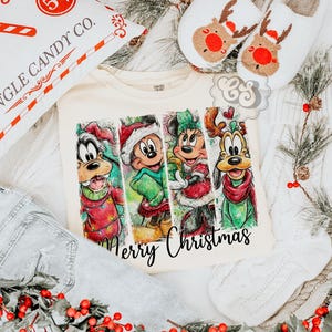 Puede incluir: Camiseta color crema con una colorida ilustraci&oacute;n de personajes de dibujos animados con atuendos navide&ntilde;os. Las palabras "Merry Christmas" est&aacute;n escritas en una fuente cursiva. La imagen tambi&eacute;n incluye una almohada decorativa, zapatillas y unos vaqueros.