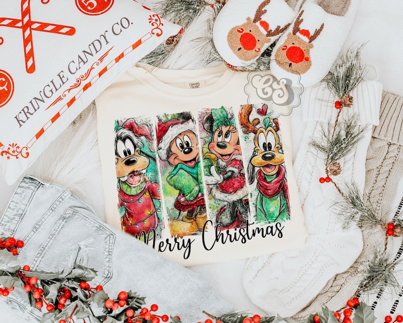 Puede incluir: Camiseta color crema con una colorida ilustraci&oacute;n de personajes de dibujos animados con atuendos navide&ntilde;os. Las palabras "Merry Christmas" est&aacute;n escritas en una fuente cursiva. La imagen tambi&eacute;n incluye una almohada decorativa, zapatillas y unos vaqueros.