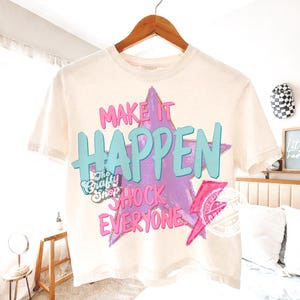 Może przedstawiać: Kremowa koszulka z napisem "MAKE IT HAPPEN SHOCK EVERYONE" w kolorze r&oacute;żowym i turkusowym, na tle fioletowej gwiazdy. Koszulka posiada r&oacute;wnież logo "The Crafty Shop" i r&oacute;żową błyskawicę.