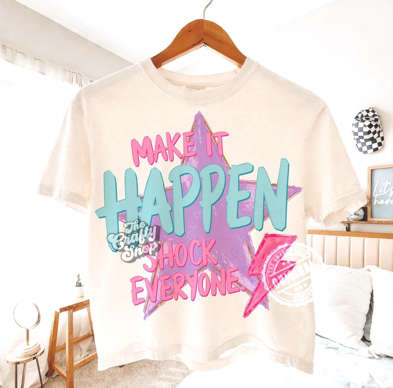 Może przedstawiać: Kremowa koszulka z napisem "MAKE IT HAPPEN SHOCK EVERYONE" w kolorze r&oacute;żowym i turkusowym, na tle fioletowej gwiazdy. Koszulka posiada r&oacute;wnież logo "The Crafty Shop" i r&oacute;żową błyskawicę.