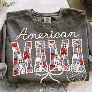 Könnte beinhalten: Ein anthrazitfarbenes Sweatshirt mit dem Schriftzug "American MAMA". Das Wort "American" ist in weißer Schrift gehalten, und "MAMA" besteht aus roten, weißen und blauen Patchwork-Elementen. Eine weiße Schleife ziert den unteren Teil des Designs.