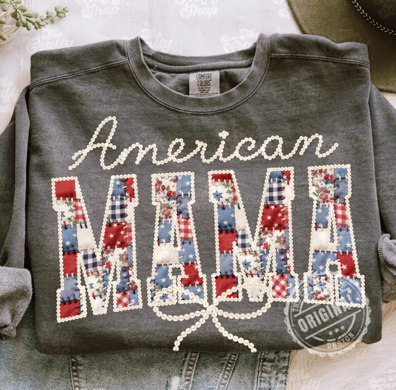 Puede incluir: Una sudadera gris oscuro con la frase "American MAMA". La palabra "American" est&aacute; escrita en blanco, y "MAMA" est&aacute; hecha de parches rojos, blancos y azules. Un lazo blanco adorna la parte inferior del dise&ntilde;o.