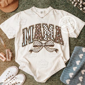 May include: A cream-coloured t-shirt featuring the word "MAMA" in a mix of patterns, including leopard print, polka dots, and checkered designs. A large bow with a checkered pattern is below the word. The shirt has short sleeves and a crew neck.