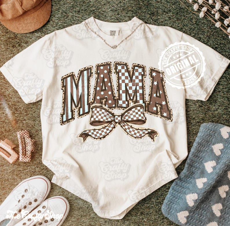 May include: A cream-coloured t-shirt featuring the word "MAMA" in a mix of patterns, including leopard print, polka dots, and checkered designs. A large bow with a checkered pattern is below the word. The shirt has short sleeves and a crew neck.