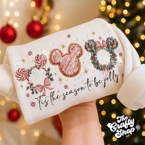 May include: A white sweatshirt featuring festive Mickey Mouse-themed designs. The design includes Mickey Mouse-shaped cookies, wreaths, and lollipops, with the phrase "Tis the season to be jolly" in script. The sweatshirt is held up against a blurred background of Christmas lights.