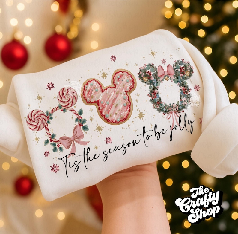 May include: A white sweatshirt featuring festive Mickey Mouse-themed designs. The design includes Mickey Mouse-shaped cookies, wreaths, and lollipops, with the phrase "Tis the season to be jolly" in script. The sweatshirt is held up against a blurred background of Christmas lights.