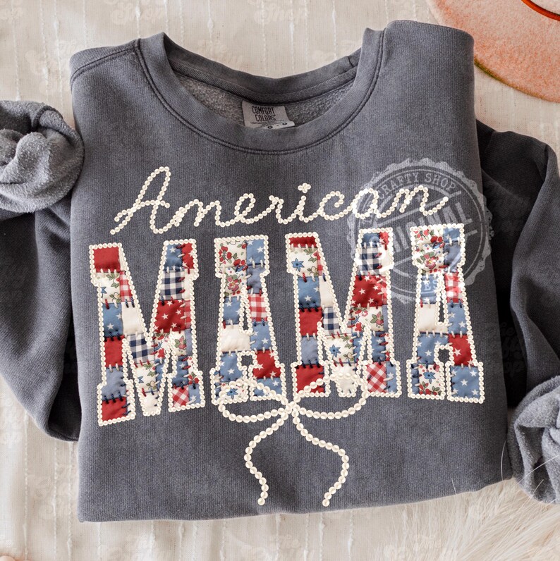 Puede incluir: Una sudadera gris con la inscripci&oacute;n "American MAMA" en letras blancas y letras de patchwork. Las letras de patchwork son rojas, blancas y azules, con un detalle de lazo debajo.