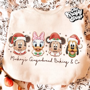 Pode incluir: Camisola creme com Mickey Mouse, Pata Daisy, Minnie Mouse e Pluto, todos usando gorros de Papai Noel. O texto "Mickey's Gingerbread Baking & Co" está impresso abaixo. O logotipo The Crafty Shop está no canto superior direito.