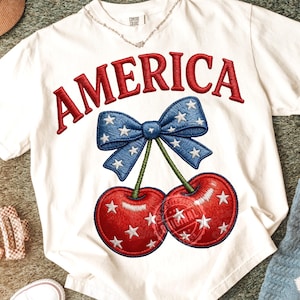 Pode incluir: Camiseta branca com a palavra "AMERICA" bordada em vermelho. Abaixo, um laço azul com estrelas brancas e duas cerejas vermelhas com estrelas brancas. A camiseta é da marca Comfort Colors.