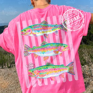 May include: A bright pink t-shirt featuring a design of three rainbow trout. The fish are vividly coloured with pink, yellow, green, and blue. Vertical white stripes are behind the fish. The text "The Crafty Shop" and "Original Design" are also visible.