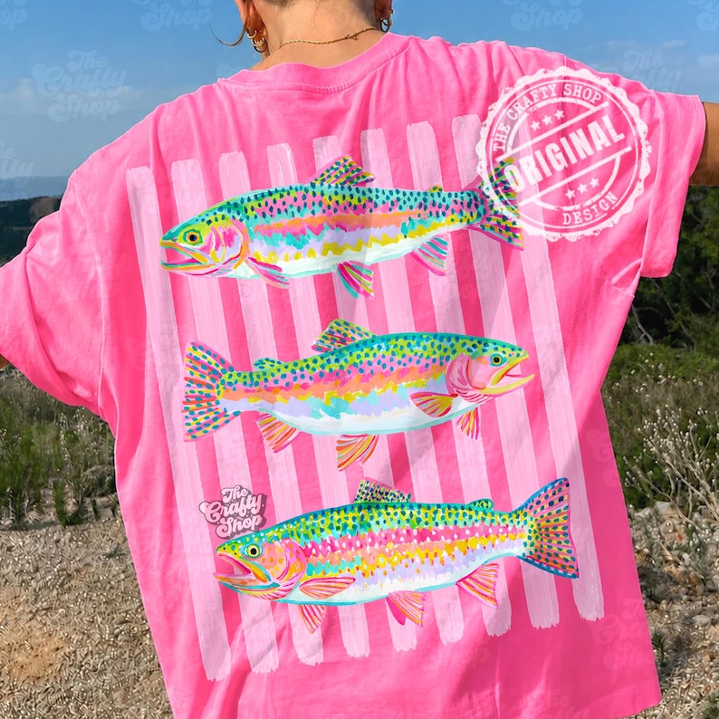May include: A bright pink t-shirt featuring a design of three rainbow trout. The fish are vividly coloured with pink, yellow, green, and blue. Vertical white stripes are behind the fish. The text "The Crafty Shop" and "Original Design" are also visible.