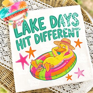 May include: A white square towel with the text "LAKE DAYS HIT DIFFERENT" in teal. A cartoon duck wearing a straw hat and floating in a colourful inner tube is featured. Pink and orange starfish accents the design. A hair clip and beaded bracelet are in the upper left corner.