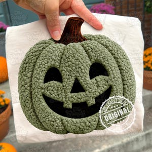 Pode incluir: Uma abóbora verde de crochê com um rosto de Jack-o'-lantern esculpido. A abóbora tem uma superfície texturizada e um caule marrom escuro. A imagem inclui um papel branco e um carimbo que diz "ORIGINAL DESIGN".