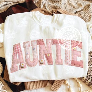 May include: White sweatshirt with the word "AUNTIE" in pink patchwork letters. The letters feature various patterns, including gingham, floral, and polka dots, with daisy accents. The sweatshirt is displayed on a neutral-toned background.