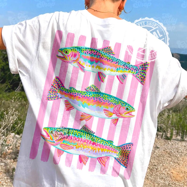 May include: White t-shirt with three rainbow trout illustrations on a pink striped background. The fish are depicted in bright colours. The shirt has a relaxed fit. The design is on the back of the shirt.