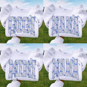May include: White sweatshirts with the words "GIGI", "MAMA", "MINI", and "MAMA" in a floral design. The floral design includes blue and pink flowers. Each word has a blue bow. The sweatshirts have puffy sleeves.