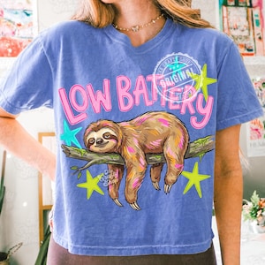 Puede incluir: Camiseta azul con un dise&ntilde;o de perezoso. El perezoso es marr&oacute;n y rosa, descansando en una rama. El texto "LOW BATTERY" est&aacute; en rosa, con estrellas amarillas y turquesas. La camiseta tiene un lavado vintage.