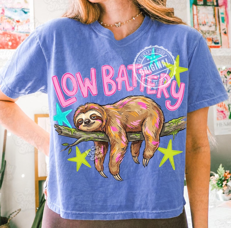 Puede incluir: Camiseta azul con un dise&ntilde;o de perezoso. El perezoso es marr&oacute;n y rosa, descansando en una rama. El texto "LOW BATTERY" est&aacute; en rosa, con estrellas amarillas y turquesas. La camiseta tiene un lavado vintage.
