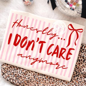 Pode incluir: Uma camiseta creme com listras verticais rosa e a frase "Honestly... I DON'T CARE anymore" em letras vermelhas. Uma fita vermelha e um selo que diz "Original" também são visíveis.