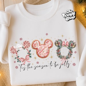 May include: White sweatshirt with a festive design featuring three Mickey Mouse head shapes. One is a candy cane wreath, one a pink cookie, and the last a floral wreath. The text "Tis the season to be jolly" is below the designs.