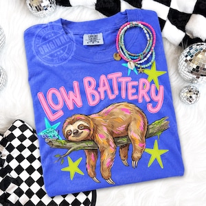 Puede incluir: Camiseta azul con la frase "LOW BATTERY" en rosa y una ilustraci&oacute;n de un perezoso. El perezoso es marr&oacute;n y descansa sobre una rama con estrellas verdes. Tambi&eacute;n se ven pulseras y bolas de discoteca.
