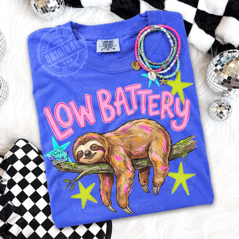 Puede incluir: Camiseta azul con la frase "LOW BATTERY" en rosa y una ilustraci&oacute;n de un perezoso. El perezoso es marr&oacute;n y descansa sobre una rama con estrellas verdes. Tambi&eacute;n se ven pulseras y bolas de discoteca.