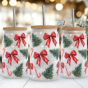 Puede incluir: Tres vasos de acero inoxidable blanco con tapa de madera y pajita de metal. Los vasos están decorados con un patrón navideño de lazos rojos, árboles de Navidad verdes y bastones de caramelo sobre un fondo blanco.