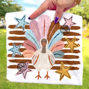 May include: A white t-shirt featuring a painted turkey graphic with a fanned tail of pastel pink, blue, and brown feathers. The design includes horizontal brown brushstrokes and five stars in blue, pink, and yellow. Two circular stamps read Original.