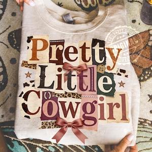 Pode incluir: Um moletom bege claro com o texto "Pretty Little Cowgirl" em estilo colagem. O texto usa várias fontes e cores, incluindo marrom, bordô e bege. O design inclui dois pequenos laços e um detalhe com estampa de leopardo.