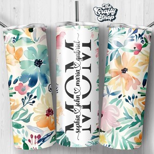 May include: A white tumbler with a floral design in shades of blue, yellow, pink, and green. The tumbler has the word "MOM" written in black with a heart and the names "sophia john maria & gabriel" written in cursive.