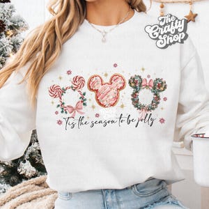 May include: White sweatshirt featuring a festive design with three Mickey Mouse-shaped elements. The design includes a candy cane wreath, a pink frosted cookie, and a floral wreath, with the text "Tis the season to be jolly."