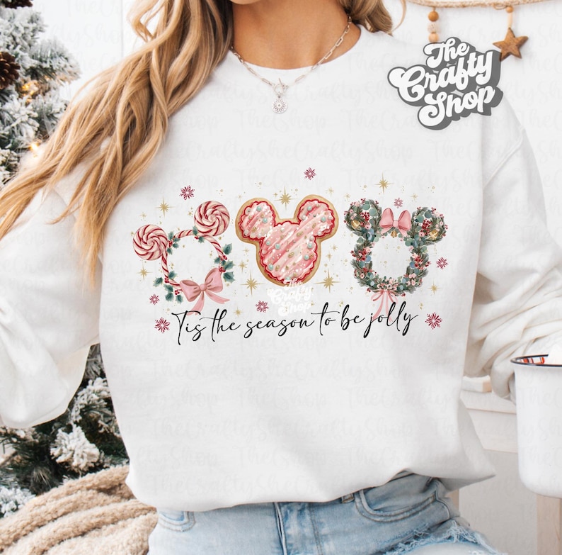 May include: White sweatshirt featuring a festive design with three Mickey Mouse-shaped elements. The design includes a candy cane wreath, a pink frosted cookie, and a floral wreath, with the text "Tis the season to be jolly."