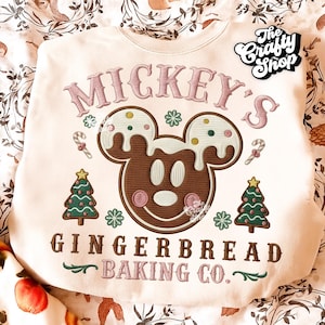 May include: Cream-colored sweatshirt with a gingerbread Mickey Mouse design. The design includes the text "Mickey's Gingerbread Baking Co." in pink and brown. The sweatshirt also features Christmas tree and candy cane graphics.