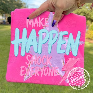 Może przedstawiać: Jasnor&oacute;żowa koszulka z napisem "MAKE IT HAPPEN SHOCK EVERYONE" w jasnoniebieskiej i r&oacute;żowej czcionce. Na koszulce znajduje się r&oacute;wnież grafika gwiazdy i błyskawicy. Koszulka jest trzymana przez dłoń.