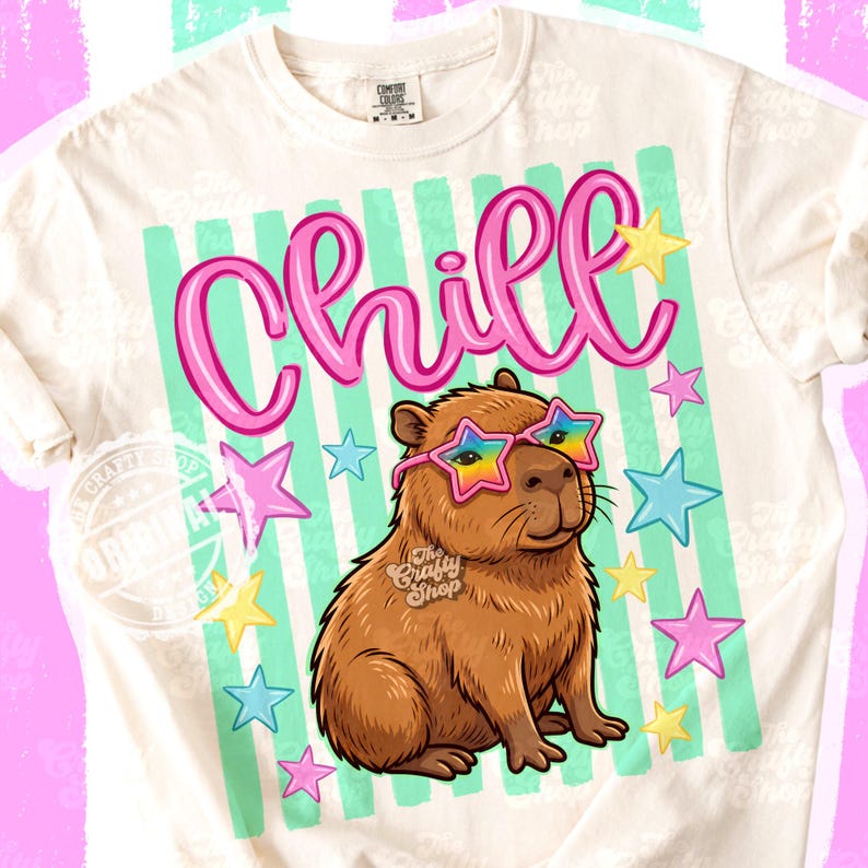 Op de afbeelding: Cr&egrave;mekleurig T-shirt met een afbeelding van een bruine capibara met een sterrenzonnebril met een regenboogontwerp. Het woord "Chill" staat in roze letters boven de capibara, met roze, gele en blauwe sterren.