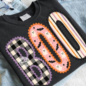 Può includere: Una t-shirt nera con la parola BOO in grandi lettere. La B è a quadri bianchi e neri, la O è rosa con fantasmi bianchi e pipistrelli neri, e la seconda O ha strisce arancioni, bianche e viola. Ogni lettera ha un bordo decorativo.