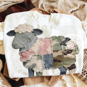 Pode incluir: Camisola branca de gola redonda com um design de ovelha em patchwork. A ovelha tem uma cabeça cinzenta, um corpo com padrão rosa, azul e camuflado e pernas azuis. O desenho está centrado na frente da camisa.