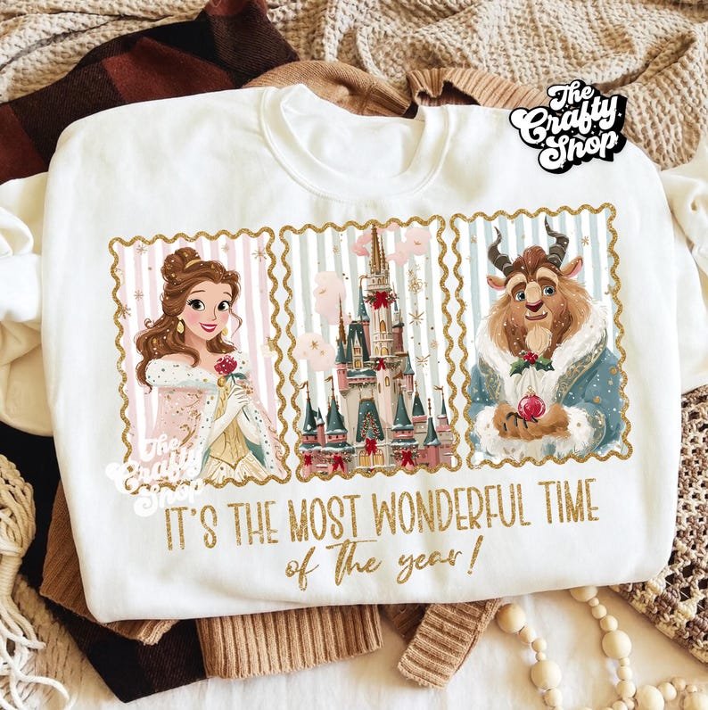Puede incluir: Sudadera blanca de cuello redondo con un gr&aacute;fico de Bella, la Bestia y un castillo, con el texto "It's The Most Wonderful Time of The Year!". El dise&ntilde;o est&aacute; enmarcado con bordes dorados y rayas rosas y azules.