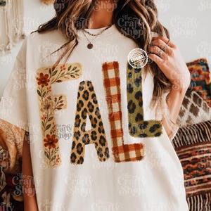 Pode incluir: Uma t-shirt cor de creme com a palavra FALL em letras grandes. O F está decorado com motivos florais, o A tem um padrão de leopardo, o L é aos quadrados vermelhos e brancos e o segundo L apresenta um padrão de leopardo verde.