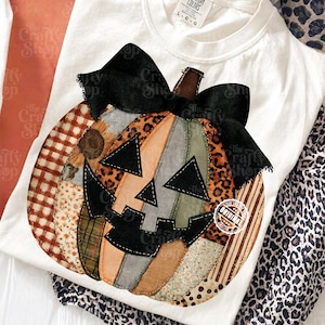 Pode incluir: Uma t-shirt branca com um gráfico grande de uma abóbora em patchwork. A abóbora é composta por vários padrões de tecido, incluindo xadrez, estampado de leopardo e motivos de girassóis, com um grande laço preto e uma cara de abóbora preta.