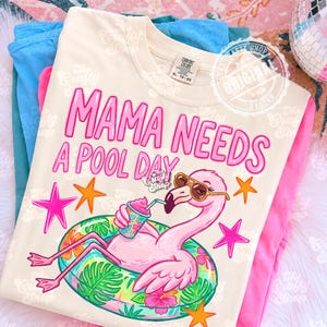 May include: Cream-colored t-shirt with the text "MAMA NEEDS A POOL DAY" in pink. The graphic features a pink flamingo wearing sunglasses and floating on a pool float, holding a drink. The shirt is folded with blue and pink clothing.