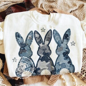 Könnte beinhalten: Weißes Sweatshirt mit Rundhalsausschnitt und drei blauen Tarnhasen-Silhouetten und gestickten Sternen. Die Hasen haben ein denimartiges Muster. Das Sweatshirt hat einen runden Ausschnitt und lange Ärmel. Das Design enthält den Text "ORIGINAL" in einem kreisförmigen Logo.